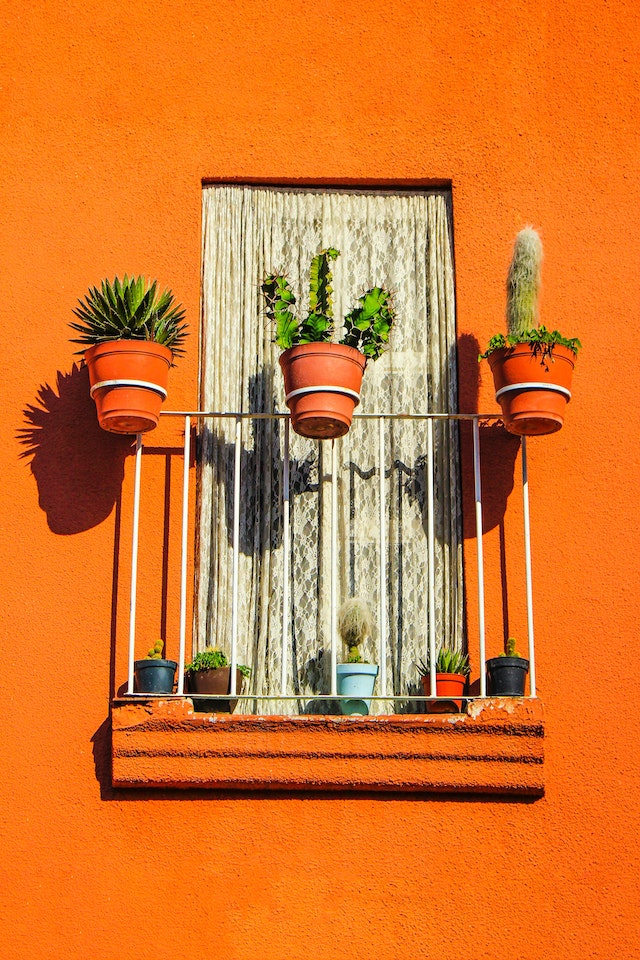 A window with a balcony railing on a house with an orange exterior. The railing holds three orange planting pots containing different succulents.